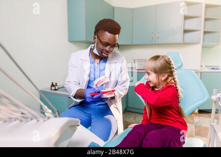 Fröhlicher lächelnder schwarzer Zahnarzt erzählt kaukasischen kleinen Mädchen, wie sie ihre Zähne putzen. Karies Prävention, Zahnheilkunde, Zahnhygiene Konzept. Stockfoto