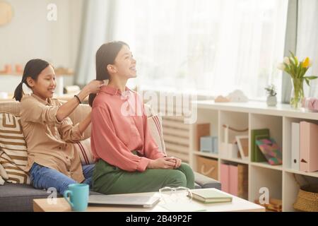 Horizontaler Schuss des glücklichen Teenager-Mädchens, das auf dem Sofa in einem gemütlichen Wohnzimmer sitzt und das Haar ihrer Mutter plaittiert, Kopierraum Stockfoto