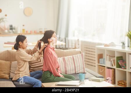 Horizontaler Seitenansicht des Teenager-Mädchens, das auf dem Sofa im gemütlichen Wohnzimmer sitzt und das Haar ihrer Mutter plaittiert Stockfoto