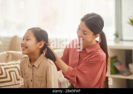 Horizontales mittleres Porträt der glücklichen Asiaten, die die Haare ihrer Tochter auf dem Sofa im Wohnzimmer sitzend machen Stockfoto