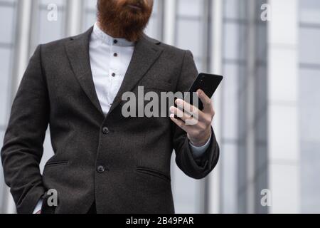 Junger bärtiger Geschäftsmann hält Handy mit App SMS-Nachricht per Jacke im Freien. Erfolgreicher Unternehmer in Form gekleidet Stockfoto