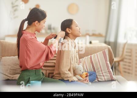 Horizontaler Seitenansicht von eleganter Asiatin, die auf dem Sofa sitzt und die Haare ihrer Tochter plaitt, Kopierraum Stockfoto
