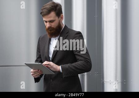 Moderner Junger bärtiger Business man arbeitet mit einem digitalen Tablet. Junge Unternehmer des Hüpfers halten Tablette in den Händen im Freien. Online mit einem arbeiten Stockfoto