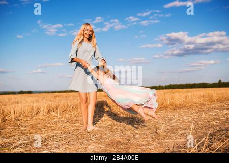 Fröhliche junge Mutter in blauem Kleid dreht sich mit ihrer kleinen schönen Tochter, die barfuß auf dem Feld steht, an einem warmen sonnigen Sommertag. Kind Stockfoto