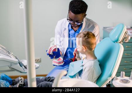 Kleines Mädchen an der Rezeption in der Zahnarztpraxis. Kleines Mädchen, das nach Zahnbehandlung und Givi in einem Stuhl in der Nähe des afroamerikanischen Zahnarztes sitzt Stockfoto