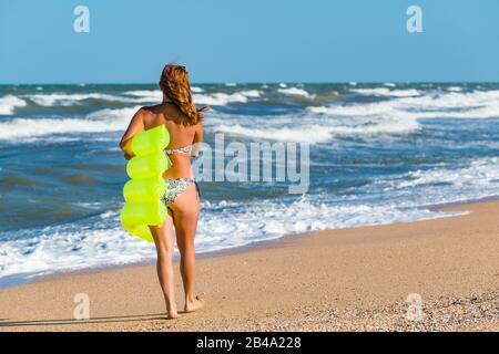 Schöne junge Frau im Badeanzug läuft am Strand entlang mit einer Luftmatratze in den Händen nahe den schaumig stürmischen Meereswellen an einem sonnigen Sommertag. Die Stockfoto