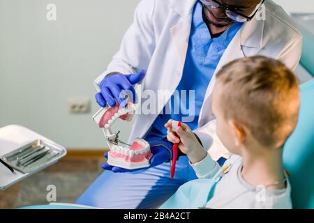 Ein lächelnder junger afroamerikanischer Zahnarzt hält künstliche Kiefer in seinen Armen, und ein kleiner Junge bürstet mit Zahnbürstenzahn-Layout. Pädiatrische Zahnmedizin Stockfoto