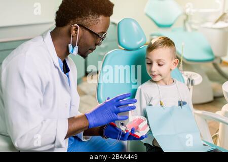 Afrikanischer amerikanischer Mann, der in einer Uniform ist. Kunststoffbacke in der Zahnarztpraxis. Dennist hält Zahnimplantate in seinen Händen und spricht mit einem kleinen Jungen Stockfoto