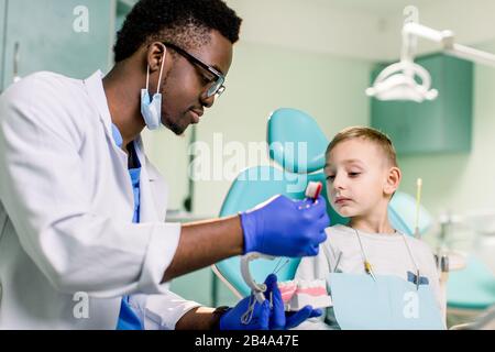 Afrikanischer amerikanischer Mann, der in einer Uniform ist. Kunststoffbacke in der Zahnarztpraxis. Dennist hält Zahnimplantate in seinen Händen und spricht mit einem kleinen Jungen Stockfoto