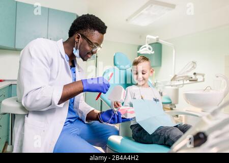 Fröhlicher lächelnder schwarzer Zahnarzt erzählt kaukasischen kleinen Jungen, wie sie ihre Zähne putzen. Karies Prävention, Zahnheilkunde, Zahnhygiene Konzept. Stockfoto