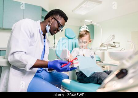 Fröhlicher lächelnder schwarzer Zahnarzt erzählt kaukasischen kleinen Jungen, wie sie ihre Zähne putzen. Karies Prävention, Zahnheilkunde, Zahnhygiene Konzept. Stockfoto