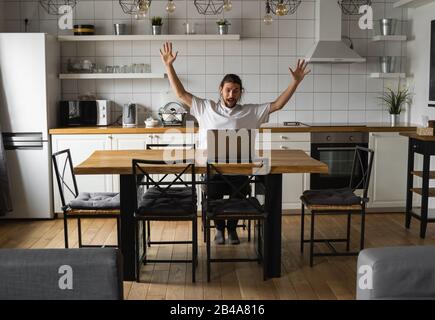 Aufgeregter Geschäftsmann freut sich über gelungenes Projekt. Mann, der die Hände in der Luft hält und auf den Laptop schaut, während er zu Hause auf einer Küche sitzt. Hap Stockfoto