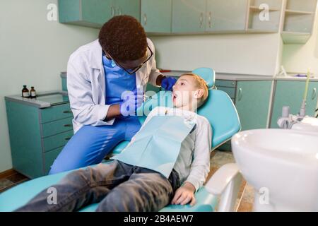 Junger schöner Junge im Zahnarztbüro. Der afroamerikanische Kinderzahnarzt untersucht die Zähne eines Kindes Stockfoto