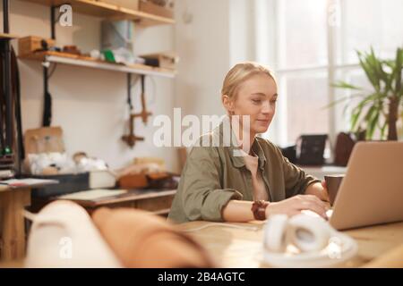 Horizontales Porträt der jungen kaukasischen Frau, die in ihrer Werkstatt mit einem Laptop beiläufige Kleidung am Holztisch trägt Stockfoto