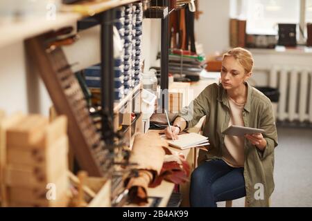 Horizontales Hochwinkelporträt der ernsthaften jungen Frau, die am Tisch in Handwerker-Worshop sitzt und Tablet-PC hält und Notizen im Notebook macht Stockfoto