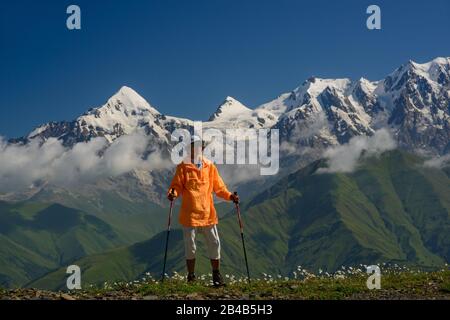 Tourist mit orangefarbener Jacke gegen Berge mit Schneehintergrund gekleidet. Alpenwiese mit weißen Blumen im Vordergrund. Stockfoto