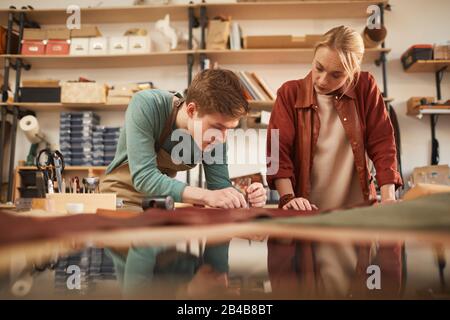 Horizontaler Schuss von zwei jungen, konzentrierten Männern und Frauen, die in der modernen Lederhandwerkswerkstatt zusammenarbeiten Stockfoto