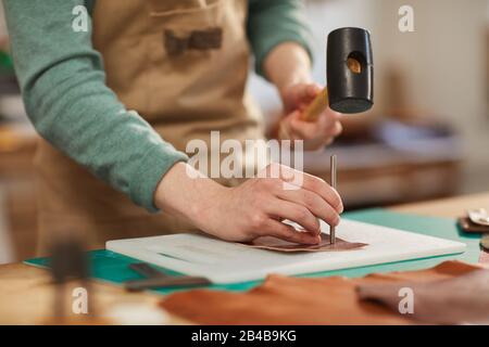 Horizontaler Nahschuss von nicht erkennbaren Handwerkern, die Schürze tragen und Löcher in Ledermaterial stanzen Stockfoto