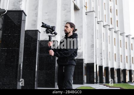 Junger professioneller Videofilmer, der professionelle Kamera auf einem 3-Achsen-Gimbal-Stabilisator hält. Die Ausrüstung hilft, hochwertige Videos ohne Shakin zu erstellen Stockfoto