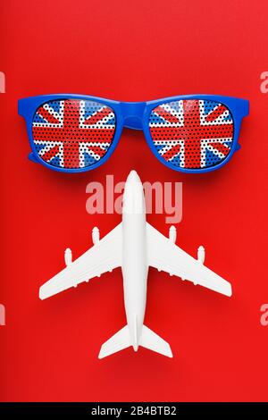 Ein weißes Passagierflugzeug fliegt in Sonnenbrille mit der Flagge Großbritanniens, auf rotem Grund. Das Konzept der Reise in England. Freier Speicherplatz. Il Stockfoto