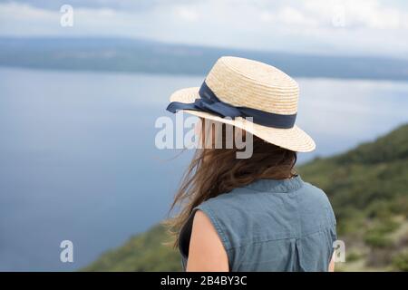 Frau in Leinenkleid und Sonnenhut steht an der Küste Kroatiens und blickt aufs Meer Stockfoto