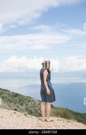 Frau in Leinenkleid und Sonnenhut steht an der Küste Kroatiens Stockfoto