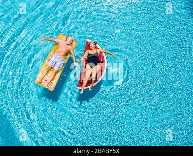 Über dem vertikalen Blick auf das alte Senioren-Paar, das sich mit Liebe in die Hände nimmt und Spaß am blauen klaren Schwimmbad hat, zusammen den Sommerurlaub mit der modischen farbigen Lilos aufblasbaren Matratze genießen Stockfoto