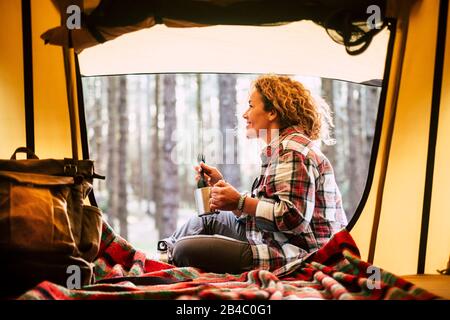 Camping mit Zelt und Abenteuer Alternative Urlaub Konzept mit fröhlichen Menschen - schöne Erwachsene blonde Lächeln und genießen Sie die Natur im Wald rund um den Ort sitzen in der Nähe eines Rucksacks und trinken Kaffee Stockfoto