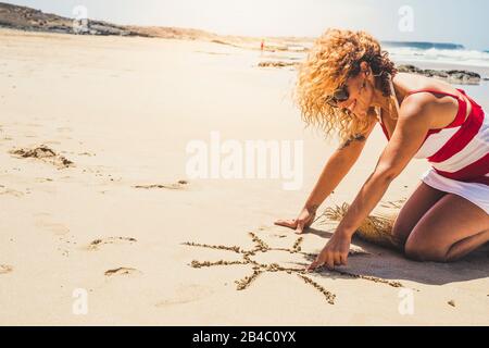 Fröhliche fröhliche Frau im Sommerurlaub am Strand und im Freien, die eine Sonne auf dem Sand gestaltet - schöne, lockige kaukasische Dame in der Freizeit im Freien Stockfoto