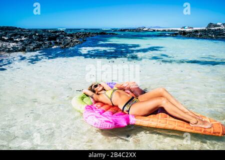 Schöne junge Frau hat ein Sonnenbad, das sich auf einer trendigen lilo aufblasbaren Matratze in einer blauen tropischen Ozeanlagune mit Sand und Felsen entspannen kann - Hafen- und Paradieskonzept für Reisen und Lifestyle Stockfoto