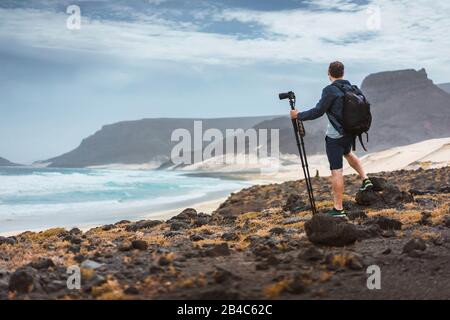 Fotograf mit Kamera auf Stativ in der Wüste, der einzigartige Landschaft von Sanddünen Vulkanklippen an der Atlantikküste einräumt. Baia das Gatas, in der Nähe von Calhau, Insel Sao Vicente, Kap Verde. Stockfoto