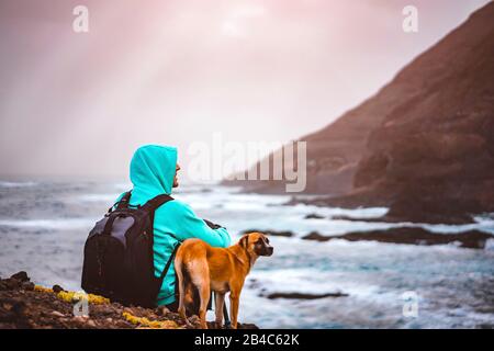 Mann mit Hund vor der ländlichen Küstenlandschaft mit Bergen und Wellen und Sonnenstrahlen, die durch die Wolken kommen. Insel Santo Antao, Kap Verde. Stockfoto