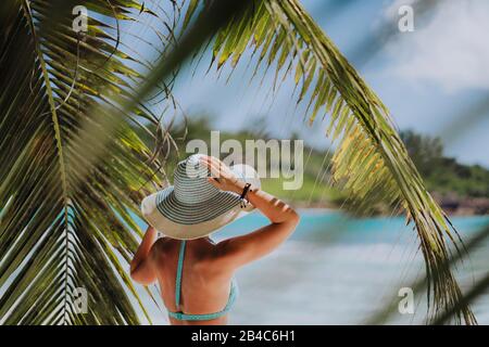 Frau am Strand im Palmenschatten mit blauem Hut. Luxus-Paradies-Erholungsurlaub Konzept. Stockfoto