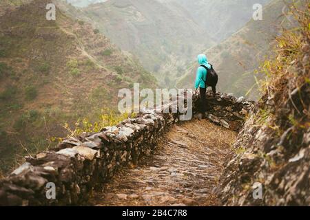 Wanderer mit Rucksack auf dem gepflasterten Pfad am Bergrand, der das Tal hinunterblickt. Felsiges Gelände mit hohen Bergketten und tiefen Schluchten um ihn herum. Santo Antao Kap Verde. Stockfoto