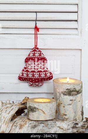 Rustikales Weihnachtsleben nordischer Stil mit Birkenkerze und niedlichem Strickweihnachtsbaum, der an einer weißen Verschlusstür hängt Stockfoto