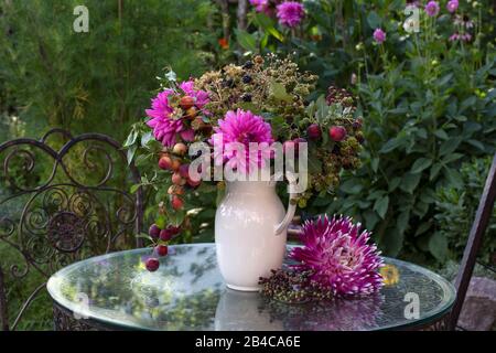 Herbststillleben mit einem Blumenstrauß aus bunten Blumen, Beeren und winzigen Äpfeln in einer weißen rustikalen Kanne, die auf einem Glastisch angeordnet ist Stockfoto