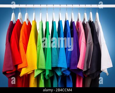 Viele neue, frische T-Shirts aus Baumwolle in bunten Regenbogenfarben hängen auf der Kleiderstange im Wrack. Verschiedene farbige Hemden auf blauweißem Backgr Stockfoto
