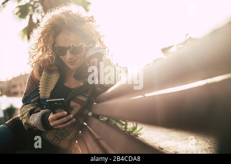 Farben in der Herbstsaison mit schöner, erwachsener, attraktiver blonder lockiger Frau verwenden ein Handy, das auf einem Beanch sitzt und bei Freizeitaktivitäten im Outoor-Bereich aktiv ist - Sonne mit Hintergrundbeleuchtung und hellem Hintergrund Stockfoto