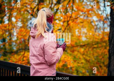 Erwachsene blonde Frau, die einen Becher in den Händen hält, steht auf einer Holzbrücke und blickt in einen Herbstwald Stockfoto