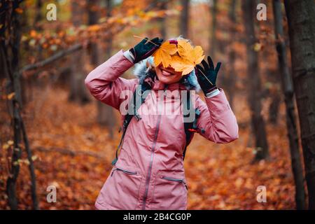 Schöne, glückliche blonde Erwachsene Frau steht mit einem Lächeln im Wald und hält gelb-orangefarbene Blätter vor ihrem Gesicht Stockfoto