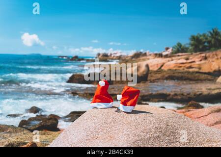 Ein Paar Weihnachtsmann hat einen tropischen, exotischen paradiesischen sandigen Strand mit Meereswellen und felsiger Küste im Hintergrund. Stockfoto