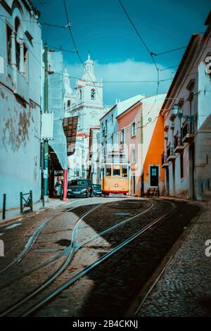 Das sommerliche Stadtbild von Lisboa. Rote Straßenbahn, die den Hügel entlang alter traditioneller Häuser hinuntergeht, sonniger Nachmittag, enge Straßen, verwinkelte Straße mit Kopfsteinpflaster, Urlaub in Lissabon. Stockfoto