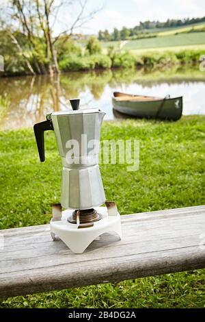 Espressotopf auf einem Campingkocher, im Hintergrund ein Kanu im Fluss Stockfoto