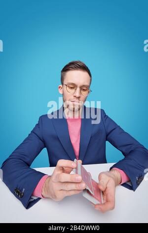 Seriöser junger Mann in Schalenform und Brille, der Spielkarten mischt, während er isoliert über blauem Hintergrund am Schreibtisch sitzt Stockfoto