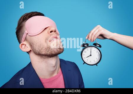 Weibliche Hand hält den Wecker mit Schlafmaske an den Augen nahe am jungen Mann, während er vor blauem Hintergrund nappt Stockfoto