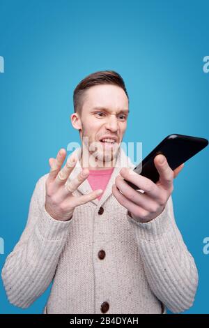 Ärgerter junger Mann mit wütender Ausdrucksweise, der auf den Smartphone-Bildschirm blickt, während er mit jemandem mit Irritationen spricht Stockfoto