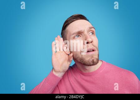 Junger bärtiger Mann in pinkfarbenem Pullover, der beim Abhören oder beim Versuch, sanften Klang zu hören, die Hand am rechten Ohr hält Stockfoto