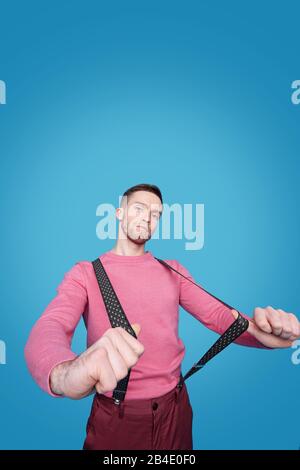 Junger gutaussehender Mann in pinkfarbenem Casual Pullover und Hose, die Hosenträger streckt, während er vor der Kamera steht Stockfoto