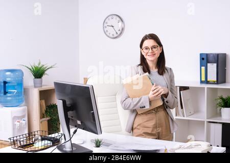 Ziemlich jung lächelnder Büroleiter oder Buchhalter mit Dokumenten, die Sie beim stehen am Arbeitsplatz mit Computer betrachten Stockfoto