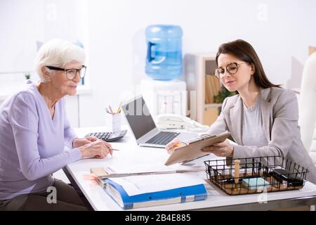 Eine elegante Frau, die auf das Versicherungsformular blickt, während junge weibliche Agentin auf einen der Punkte zeigt und es erklärt Stockfoto